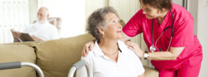 A smiling nurse crouched down, speaking to a senior woman in a wheelchair while a senior man is reading in the background