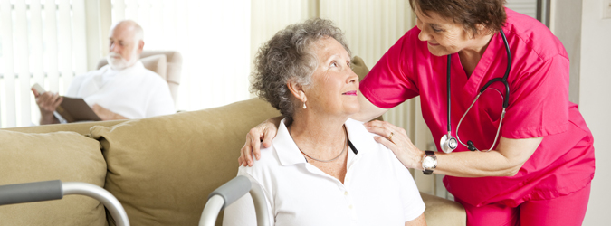 A smiling nurse crouched down, speaking to a senior woman in a wheelchair while a senior man is reading in the background