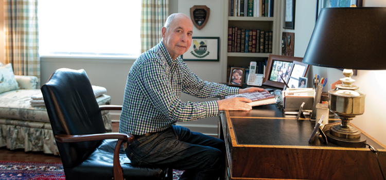 A senior man seated at a desk in a study, complete with comfortable seating and a bookshelf