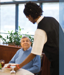 A young man serving dinner to a senior man in Whitney Center's Center Stage Restaurant