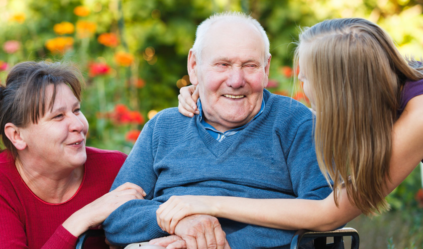 Two younger women crouched down, speaking to a senior man in a wheelchair