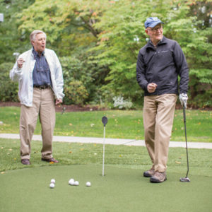 Two senior men in fall seasonal clothing practice putting on the Whitney Center putting green