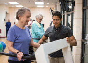 A young male staff member assists a smiling senior woman on the treadmill in the Whitney Center fitness center