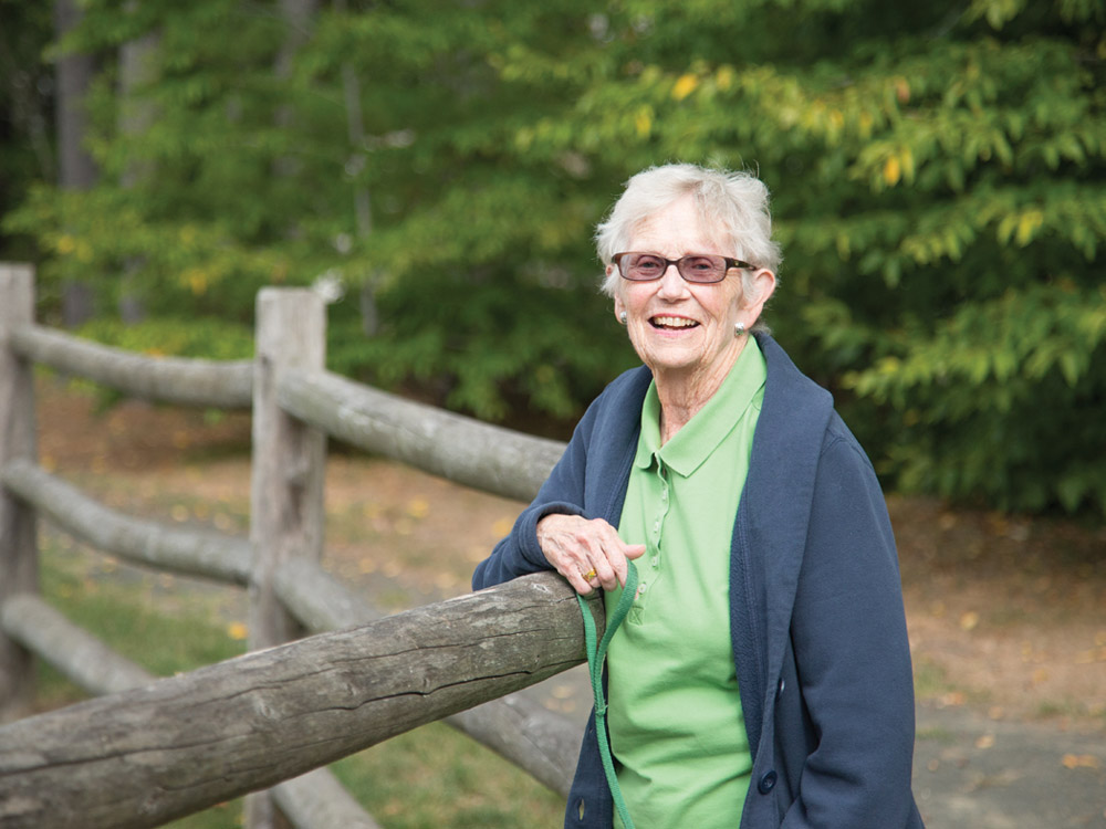 A smiling senior woman leans against a wooden railing on one of Whitney Center's on-campus walking trails