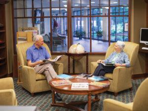 A smiling senior man and woman sitting in armchairs have a conversation in one of Whitney Center's common rooms