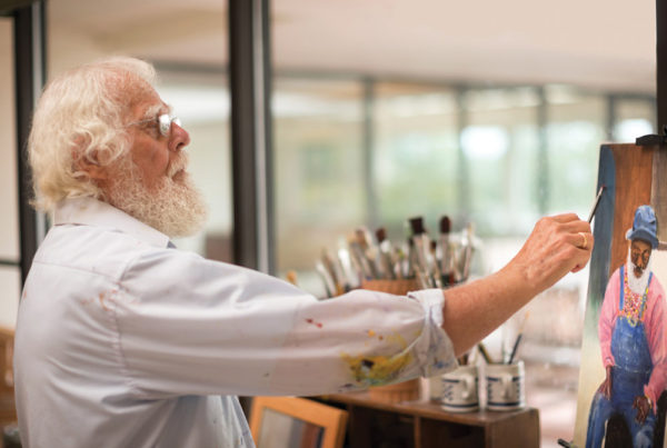 A senior man with white hair and a white beard puts the finishing touches on a painting he has created