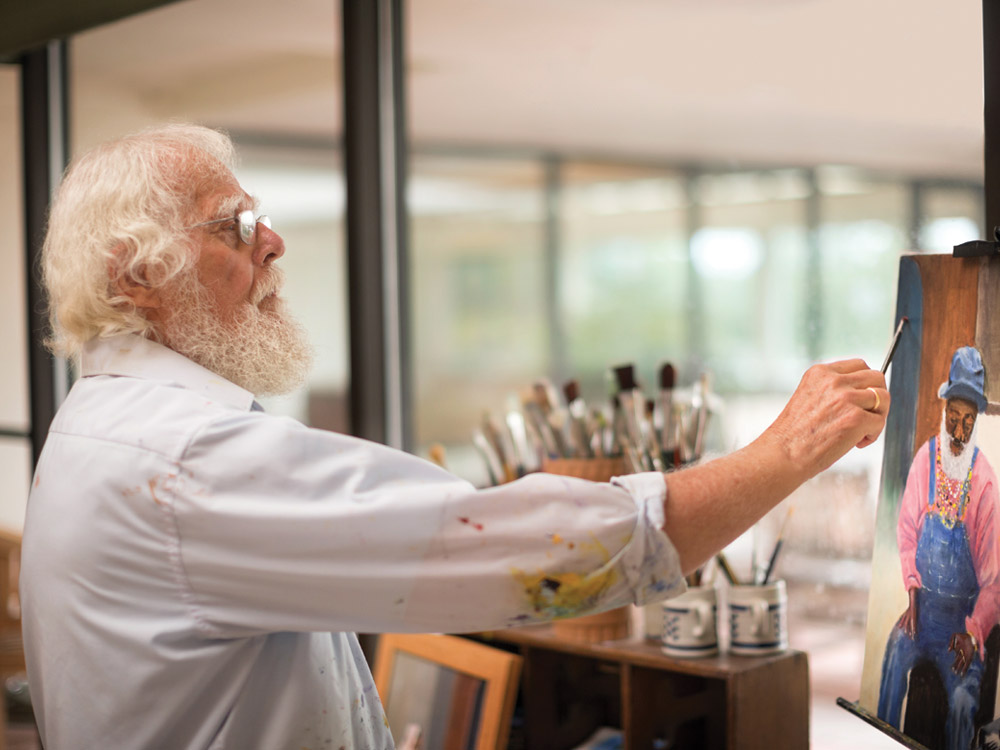 A senior man with white hair and a white beard puts the finishing touches on a painting he has created