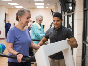 A young male staff member assists a smiling senior woman on the treadmill in the Whitney Center fitness center