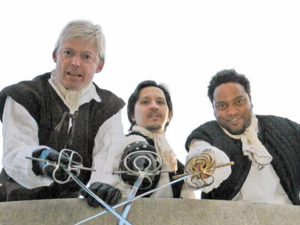 Three male actors in Shakespearean costumes cross swords while posing for a photo